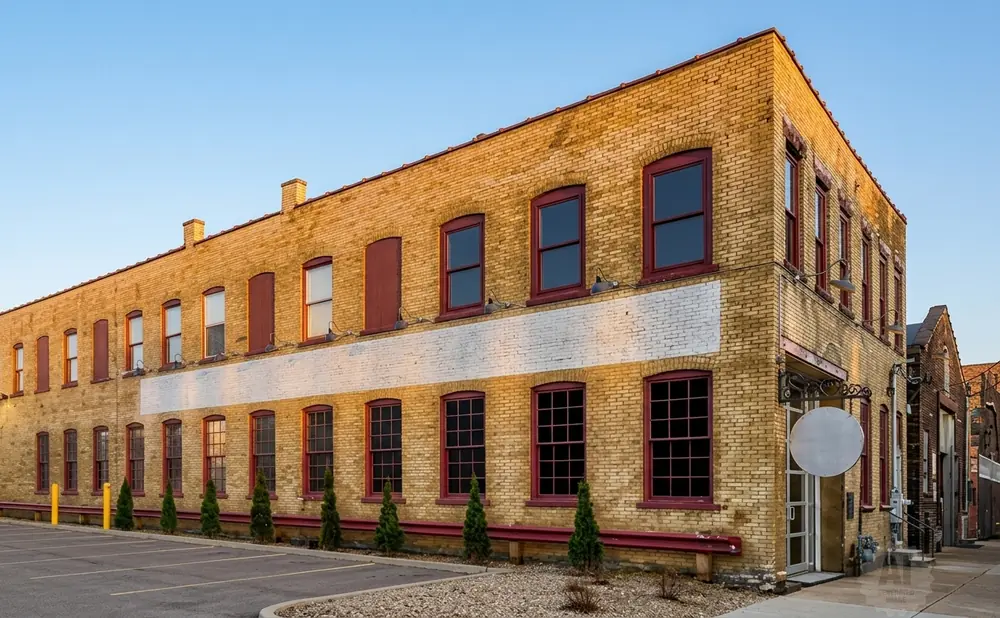 Exterior of a tan brick building with red-trimmed windows, a paved parking lot, and a clear blue sky.