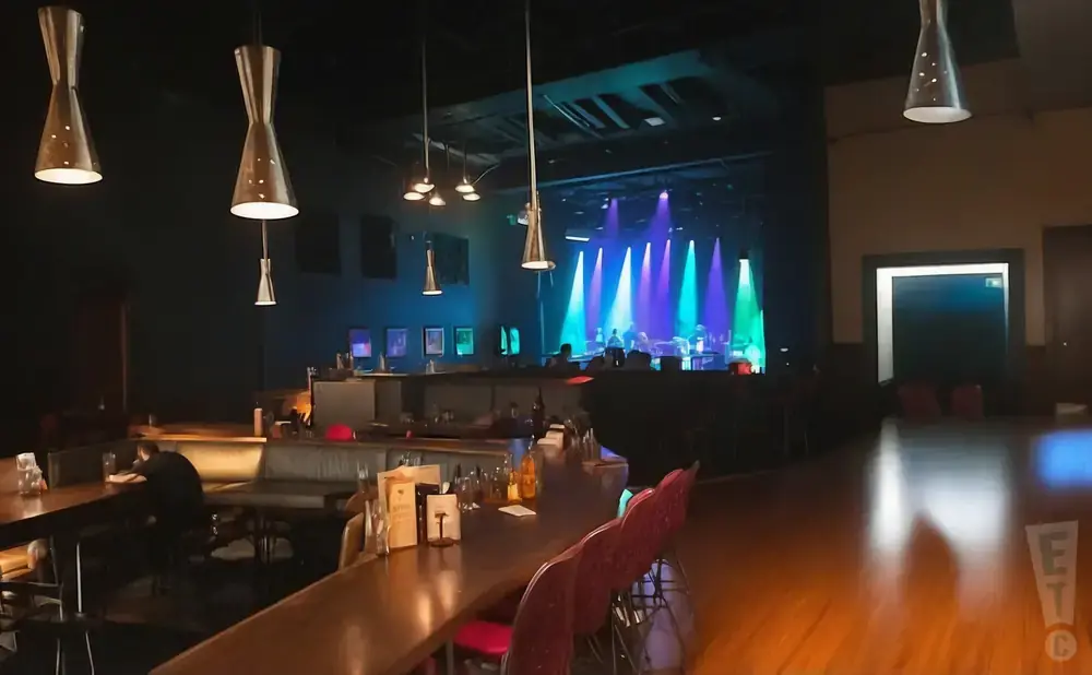 A dimly lit concert venue with a stage set up for a band, and a bar area in the foreground.