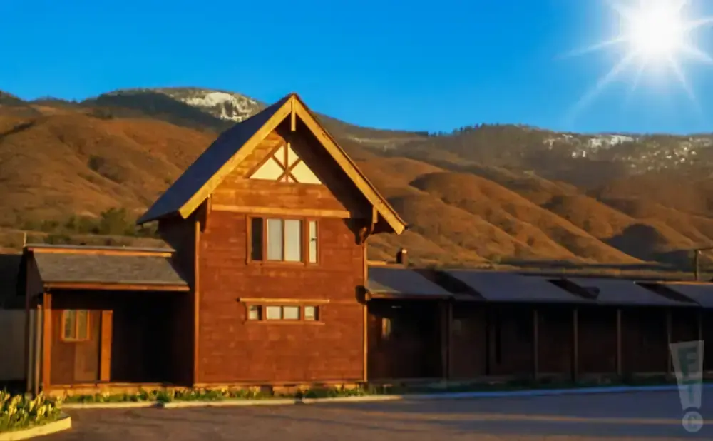 Wooden lodge with snow-capped mountains and a bright sun in a clear blue sky.