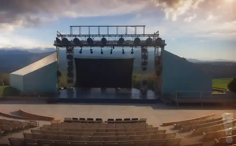 Outdoor amphitheater with stage and seating in front of a mountain landscape.