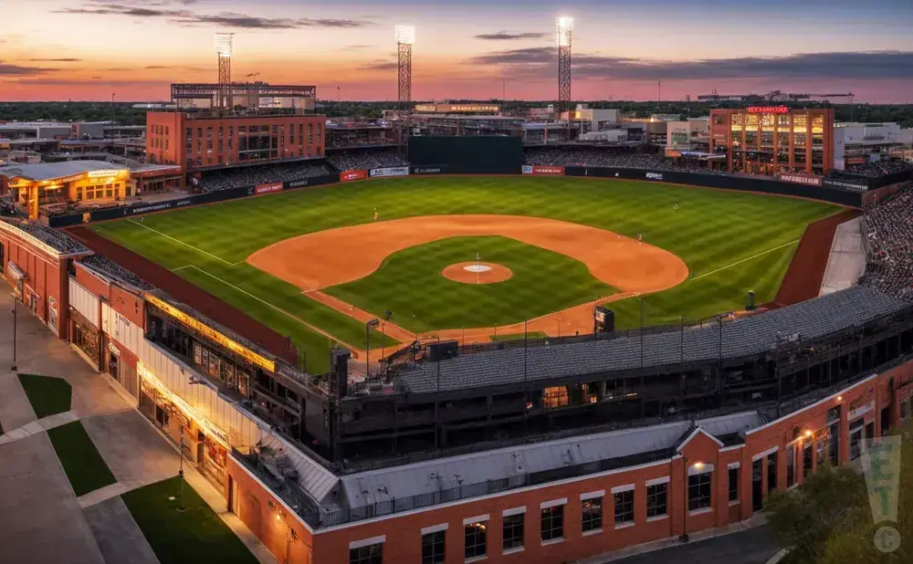 Aerial view of a baseball stadium at sunset, with the field illuminated by lights.