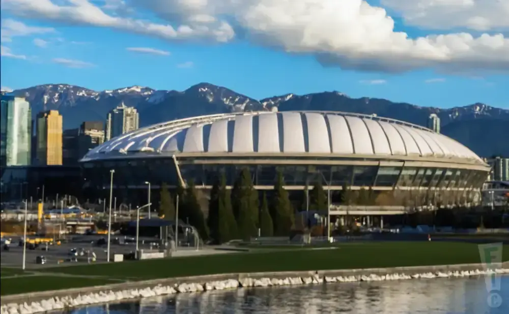 A domed stadium sits behind a waterfront, with mountains in the background under a blue sky with clouds.