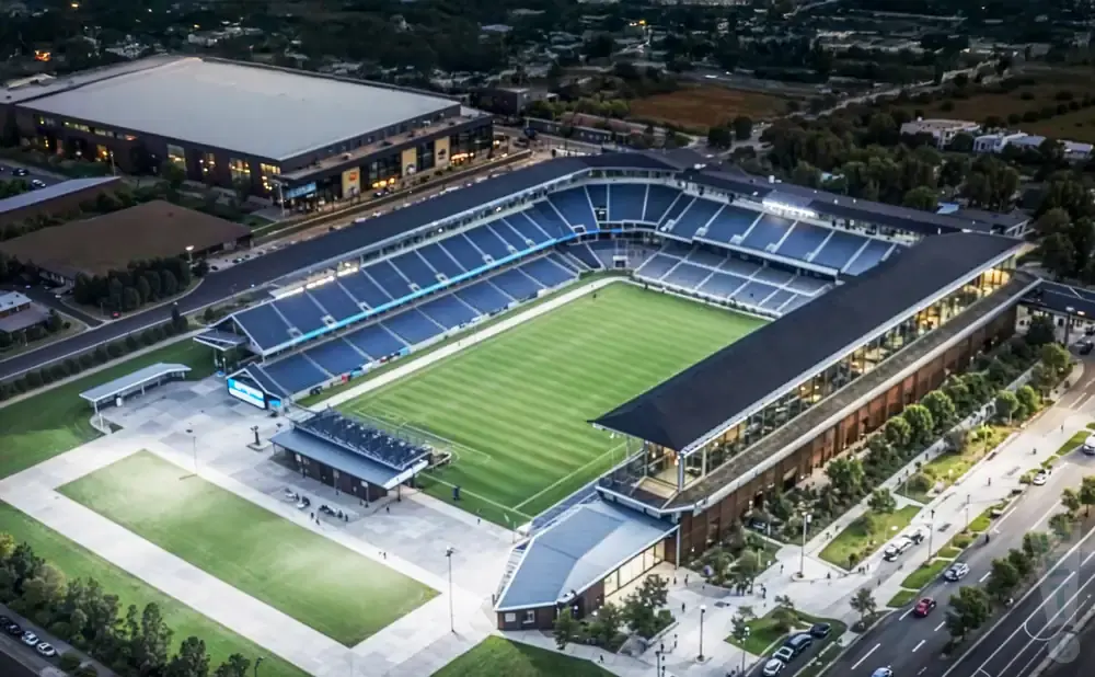 Aerial view of a soccer stadium with blue seats, a green field, and surrounding buildings and trees.
