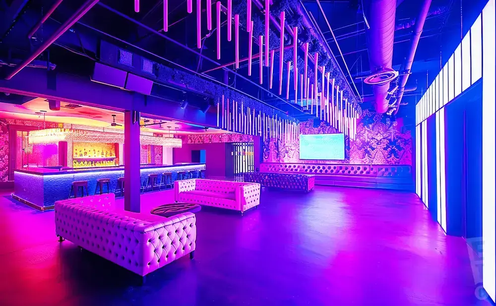 A nightclub interior with purple and pink lighting, a bar, sofas, and a large screen.