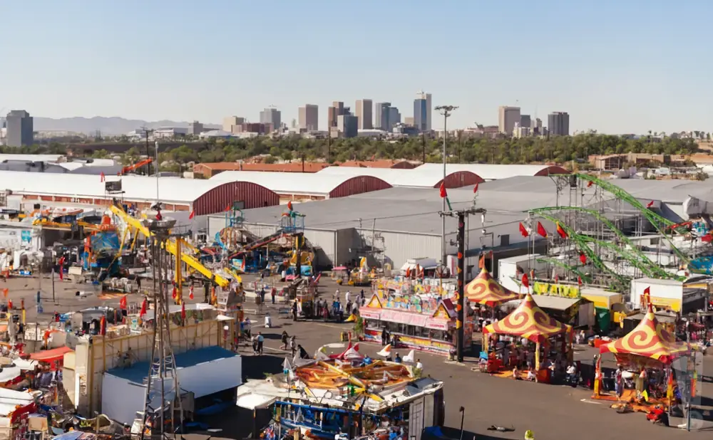 An aerial view of a bustling outdoor carnival with rides, food stands, and a city skyline in the background.