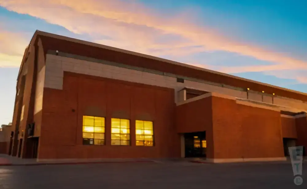 A modern brick building with large windows illuminated from within, set against a sky with soft pink and orange clouds.
