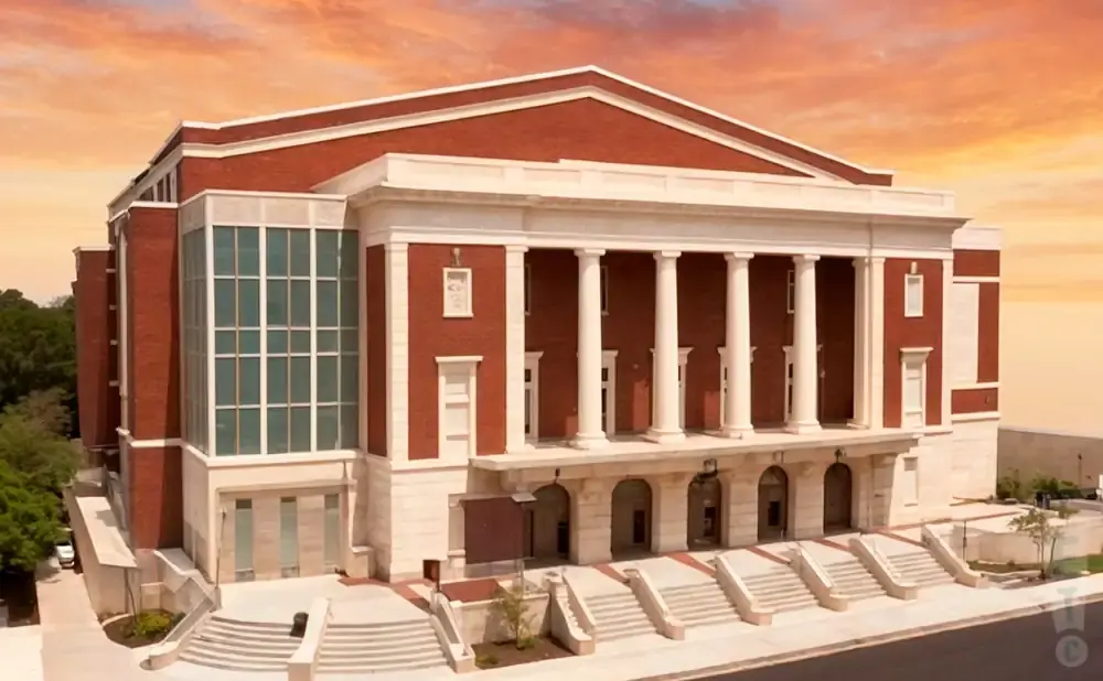 A red brick building with white columns and large windows, set against a colorful sky.