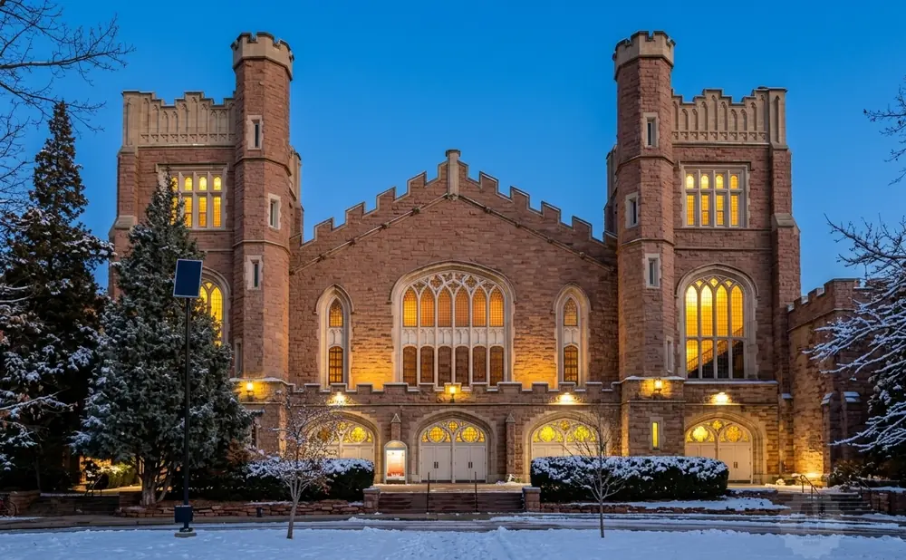 A gothic-style stone building with two towers and arched windows at dusk, surrounded by snow-covered trees.