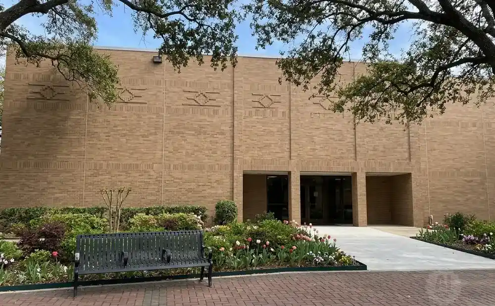 A brick building with patterned accents, a dark bench, and a flowerbed in front.