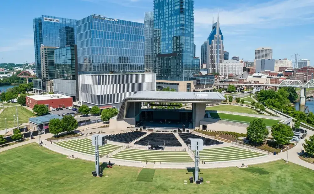 Aerial view of a modern amphitheater with tiered seating and a stage, backed by a city skyline with glass skyscrapers and a river.