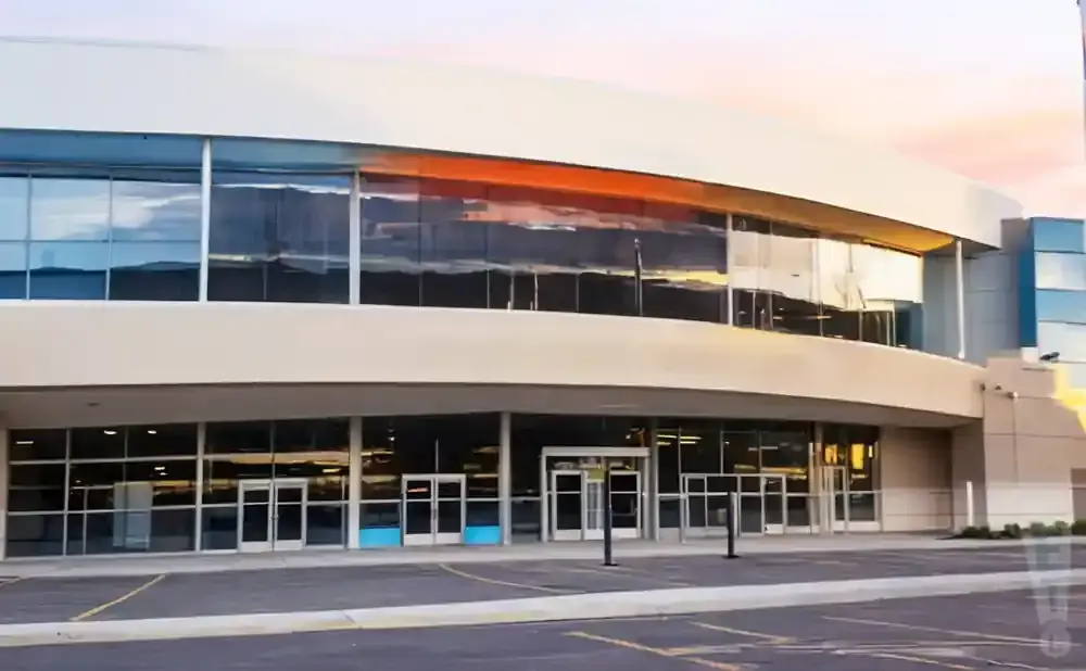 Exterior of a modern building with large glass windows reflecting a sunset, with parking lot in foreground.
