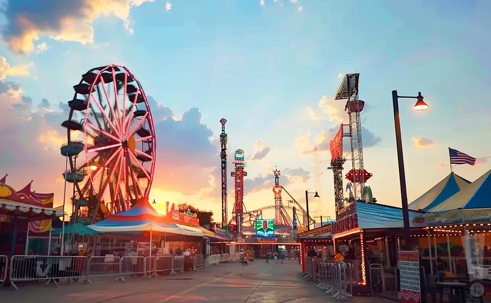 Ferris wheel and rides at a carnival at sunset, with colorful lights and a cloudy sky.