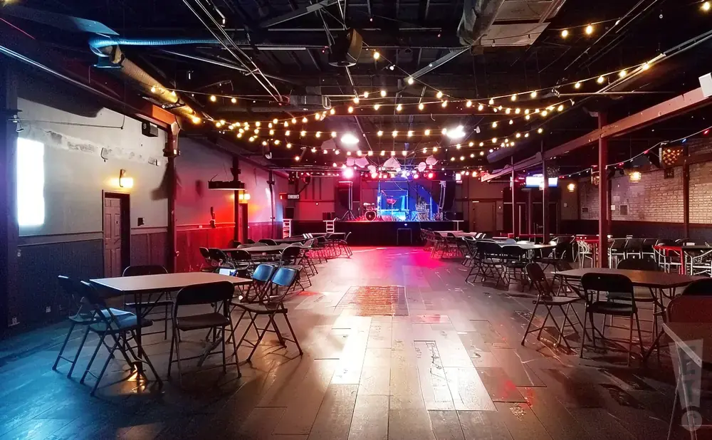 An empty concert hall with a stage, tables, and chairs, decorated with string lights.