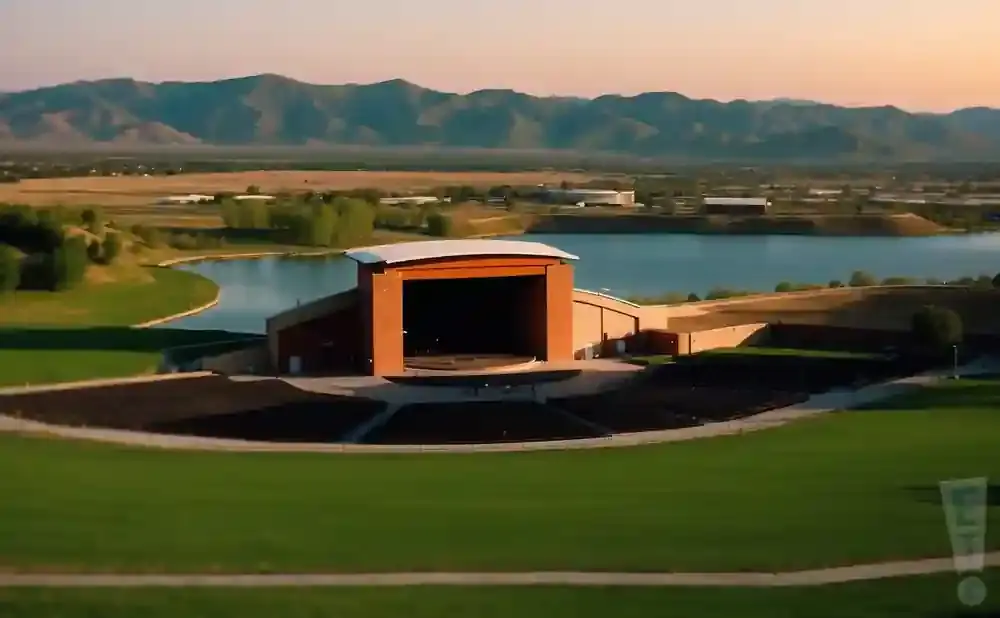 An outdoor amphitheater with a stage sits beside a lake, with mountains in the background.