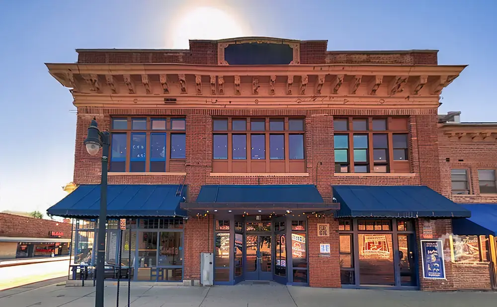 Exterior of a brick building with blue awnings and large windows, featuring a lit street lamp in front.