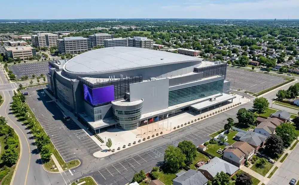 Aerial view of a large modern arena with a curved roof and purple digital screens, surrounded by parking lots and a suburban neighborhood.