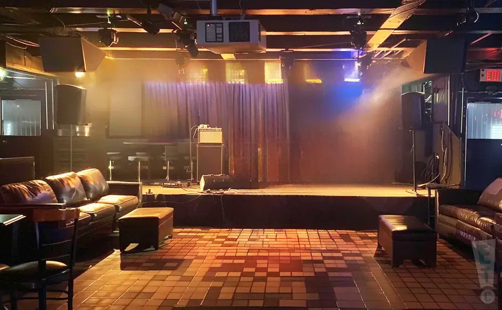 Stage and seating area in a dimly lit club with a leather couch and ottomans on a tiled floor.