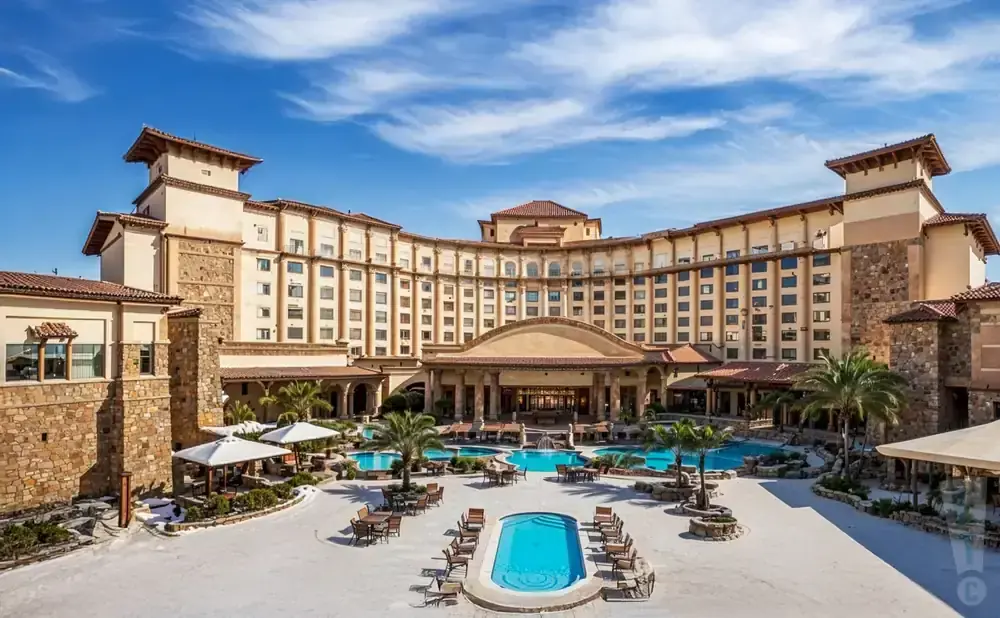 Large resort with multiple pools, palm trees, and a curved, multi-story building under a blue sky.