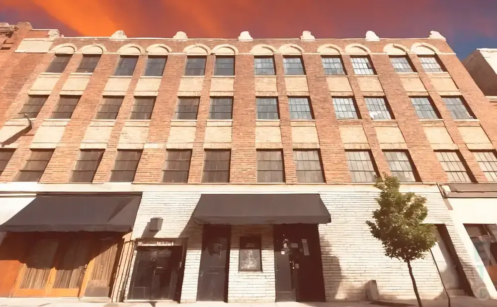 Red brick building with arched windows and awnings over storefronts. A small tree stands in front.