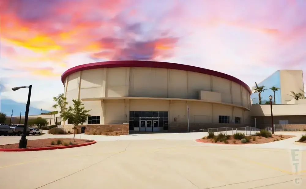 Round, tan building with a red roof, palm trees, and a colorful sunset sky.