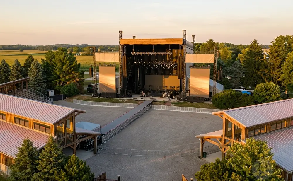 An outdoor concert stage with large screens, seating, and surrounding trees at sunset.