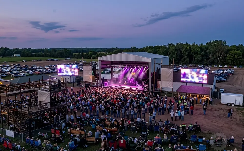 An outdoor concert at dusk with a band on stage, a large crowd, and a parking lot full of cars.