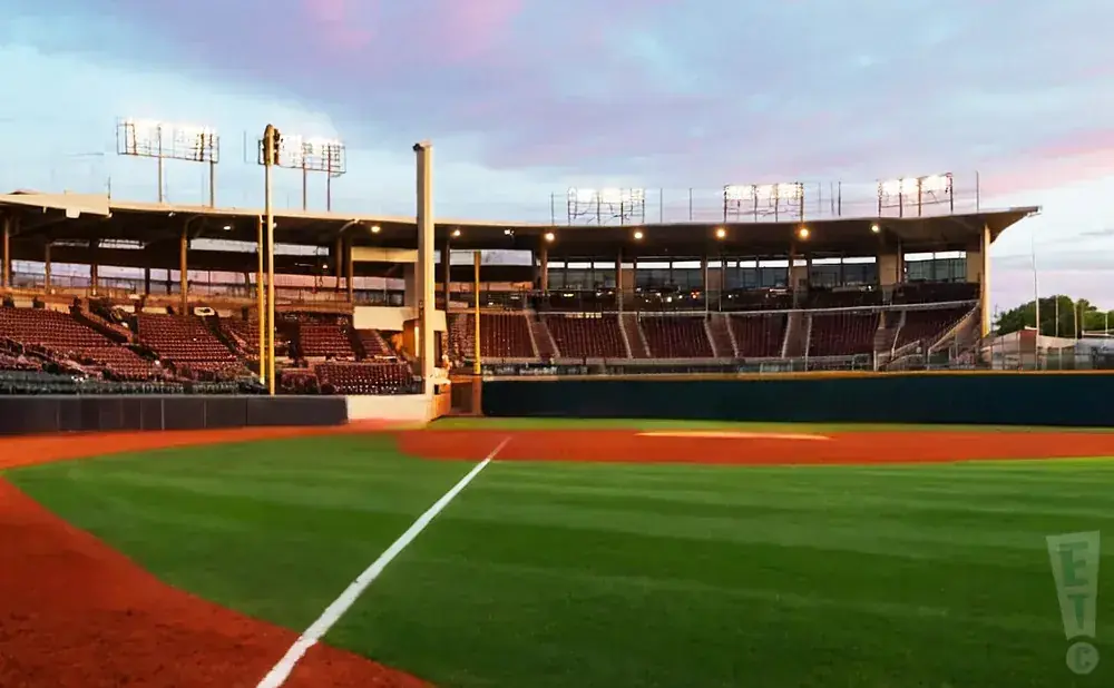 Empty baseball stadium with green grass infield and red dirt base paths, lit by stadium lights under a colorful dusk sky.