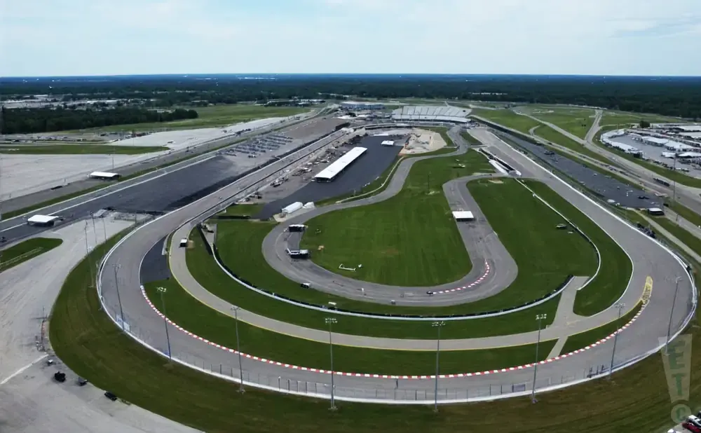 Aerial view of a large oval-shaped racetrack with grandstands, green infields, and surrounding roads and parking areas.