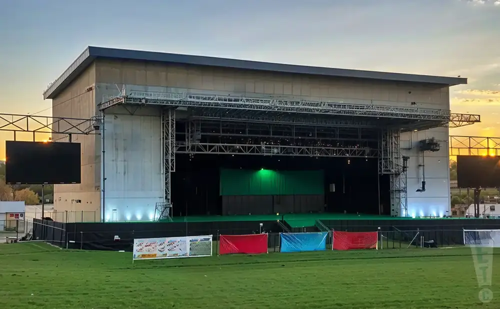 Outdoor concert stage with large screens and banners in front of a green field at sunset.