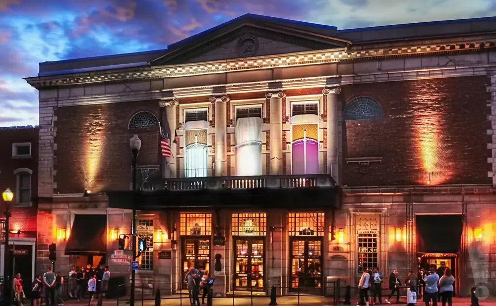 Exterior of an ornate brick building with large windows and classical columns, lit up at dusk.