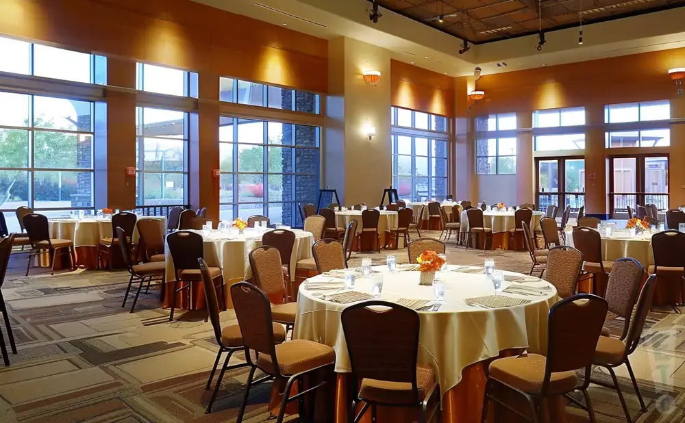 A banquet hall with round tables set for an event, large windows, and warm lighting.