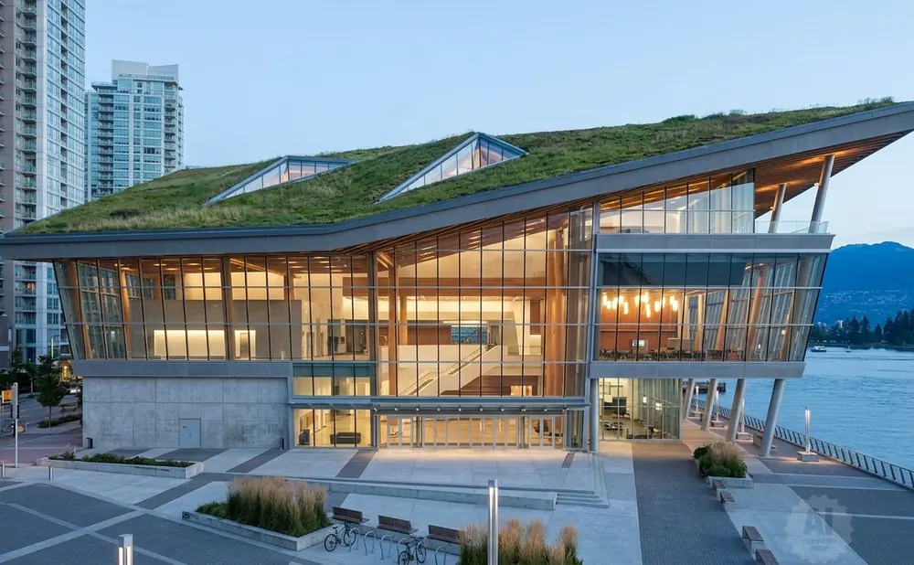 Modern building with a green roof by the water, featuring large glass windows and wooden beams.