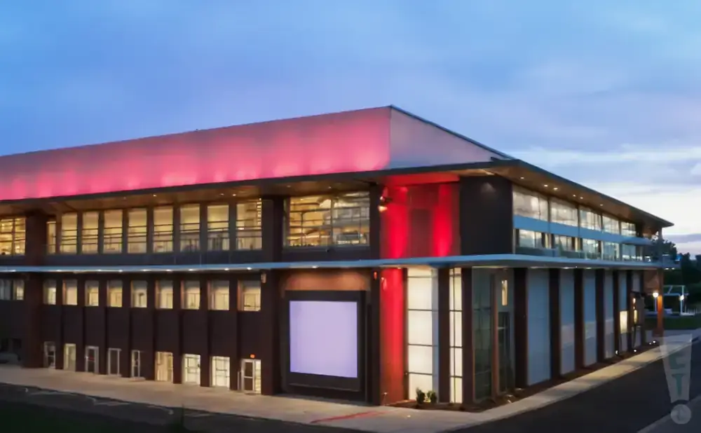 Modern brick building with red lighting at dusk, featuring large windows and a lit-up screen.