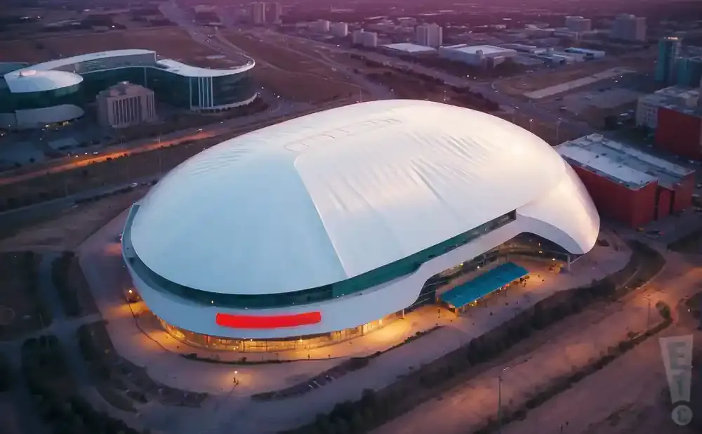 Aerial view of a large domed stadium with a white roof and a red sign, surrounded by modern buildings and city lights.