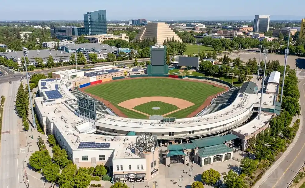 Aerial view of a baseball stadium in a city with tall buildings, a pyramid structure, and a golden bridge.