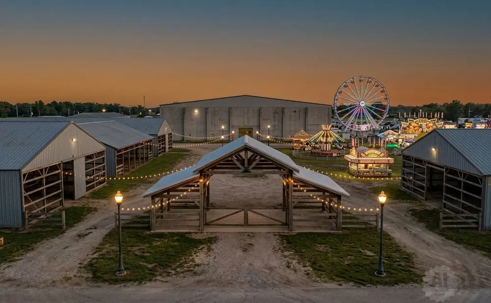 Aerial view of a fairground at dusk, featuring barns, a wooden archway, and carnival rides like a Ferris wheel.