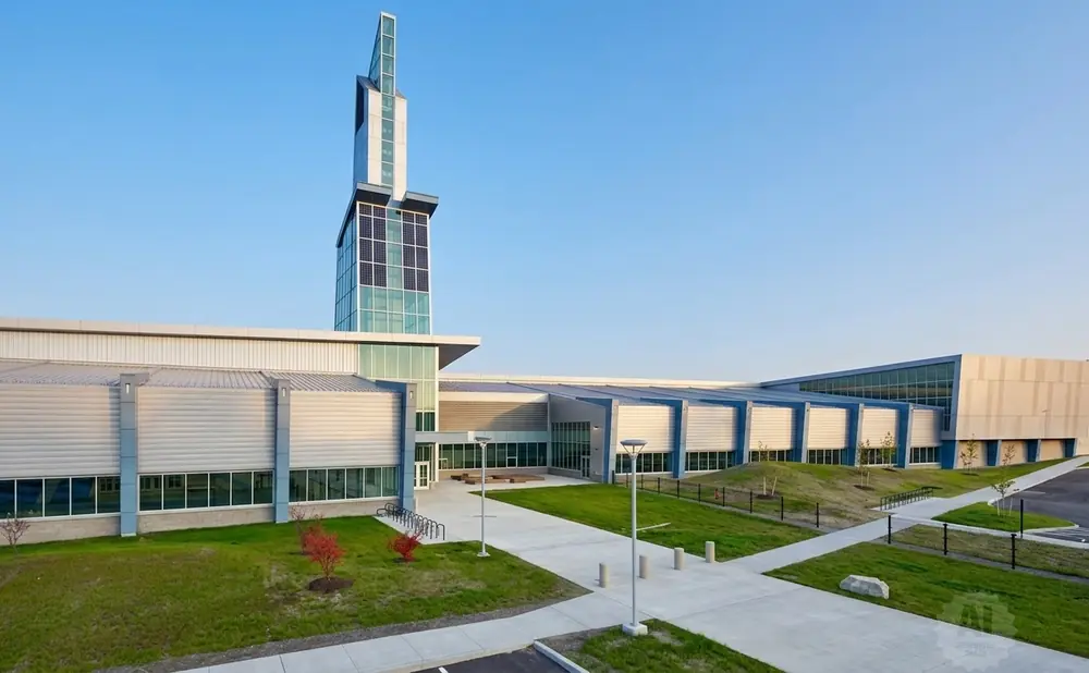 Modern building with a tall glass tower, silver siding, and large windows, surrounded by green grass and a clear blue sky.