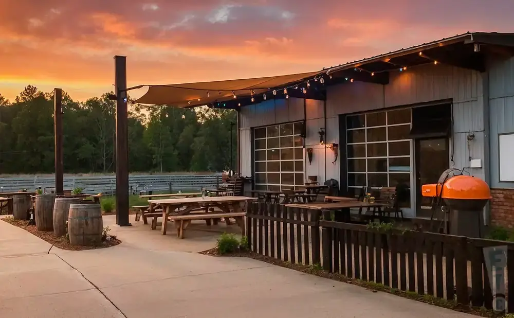 Outdoor seating area with picnic tables, barrels, and a grill under a sunset sky.