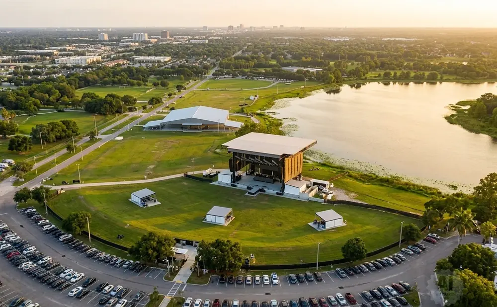 Aerial view of an outdoor amphitheater stage, lake, and parking lot at sunset.