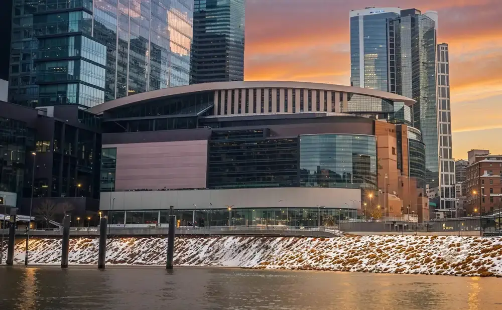 Modern arena on a riverfront with snow-dusted banks, framed by tall glass buildings and a colorful sunset sky.