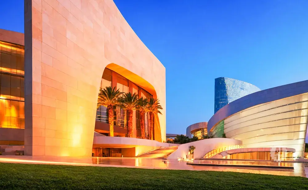 Modern architecture with curving glass and stone buildings, illuminated at dusk with palm trees.