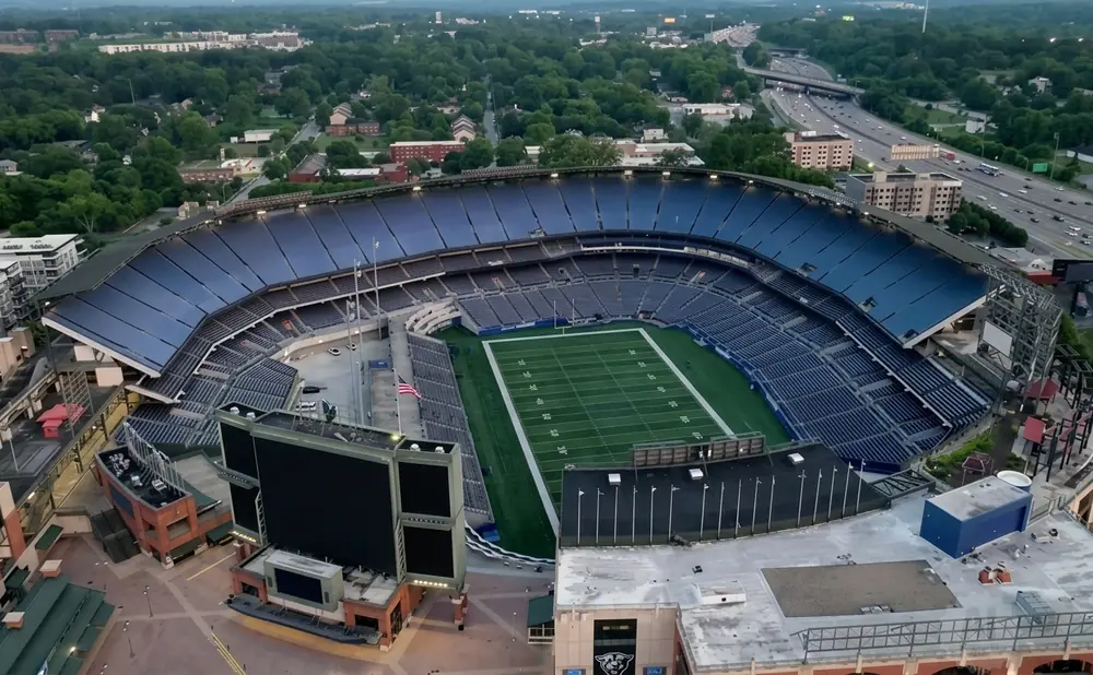 Aerial view of a football stadium with a green field and blue seats, surrounded by trees and a highway.