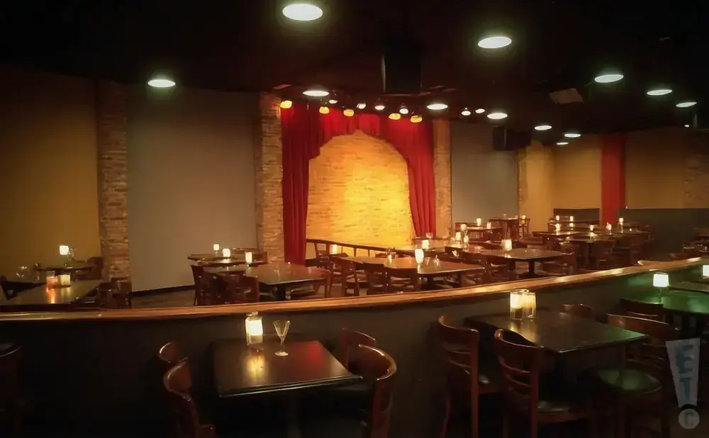 A comedy club or small theater with tables and chairs set up in front of a stage with red curtains.