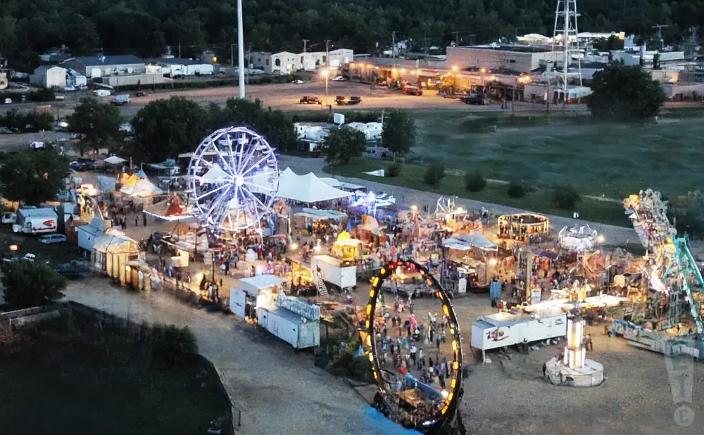 An aerial view of a brightly lit carnival with a Ferris wheel, rides, and tents filled with people.