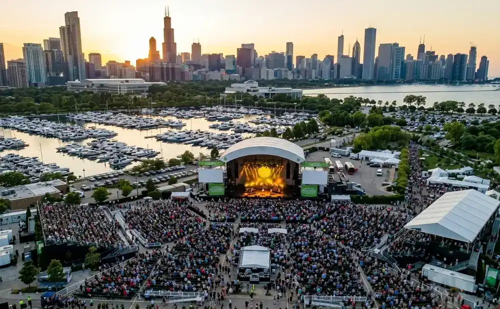 An aerial view of a concert venue with a large crowd gathered for a performance against a city skyline at sunset.