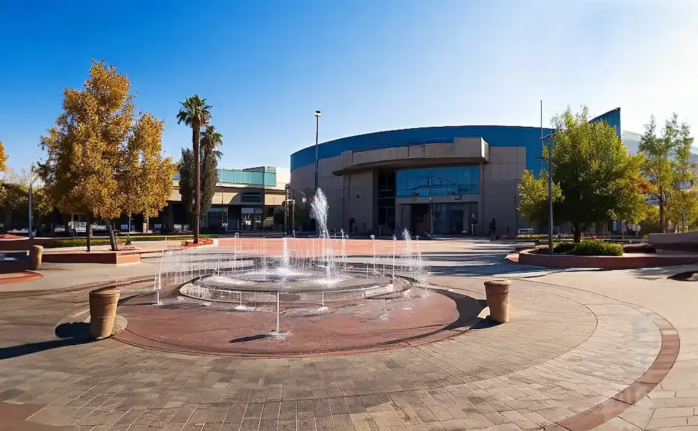 A circular fountain sprays water in front of a modern building with large windows, surrounded by trees and paved walkways.