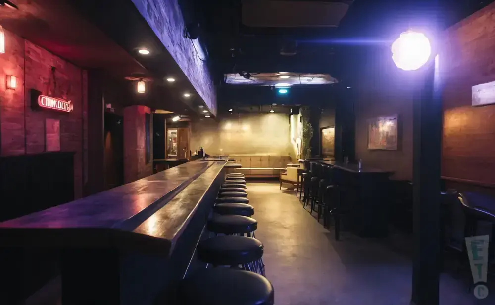 A dimly lit bar with a long counter, stools, and lounge seating in the background.