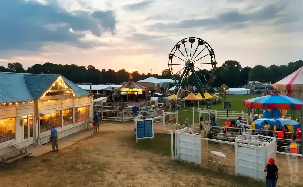An outdoor fairground with rides, games, and food stands at sunset, with a Ferris wheel and a carousel.