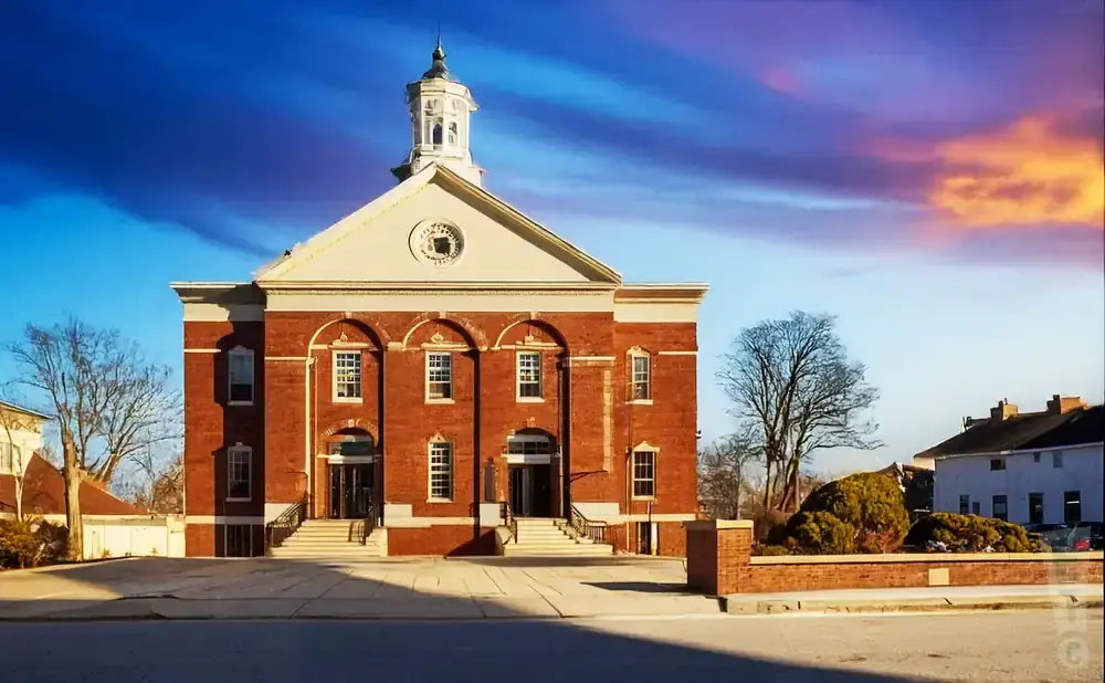 A brick building with a white cupola stands against a dramatic sunset sky.