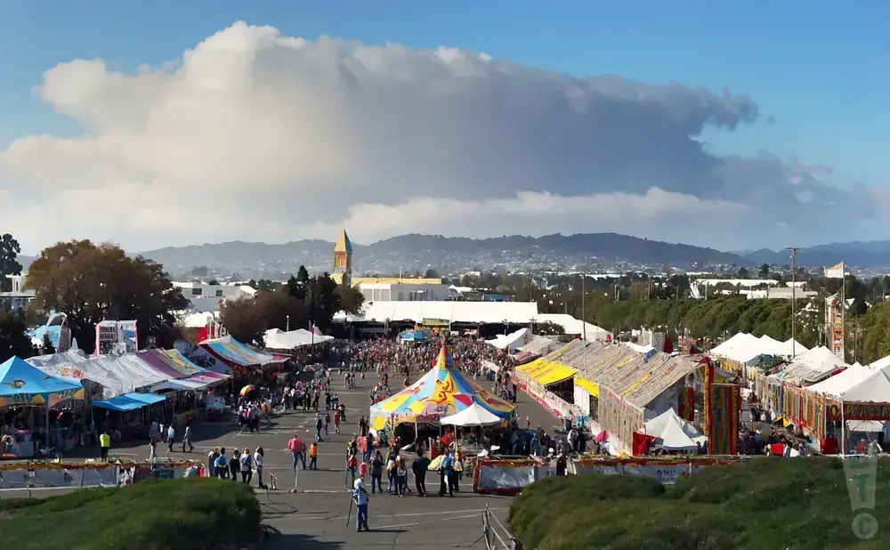 Crowded outdoor fair with colorful tents, food stalls, and distant buildings under a cloudy sky.